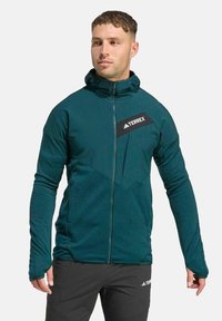 Grüne Terrex-Jacke mit durchgehendem Reißverschluss, hohem Kragen und langen Ärmeln. Verfügt über ein diagonales schwarzes Logo-Patch auf der Brust und eine strukturierte Stoffoberfläche.