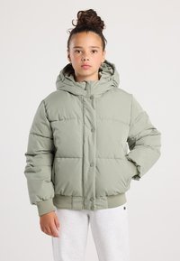 Joven con cabello rizado recogido en un moño, vistiendo una chaqueta acolchada de color verde claro con capucha y pantalones de chándal gris claro, de pie frente a un fondo liso.