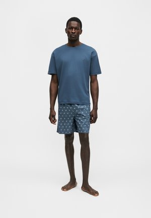 Homme debout pieds nus portant une chemise bleue à manches courtes et un short bleu à motifs sur un fond blanc uni.