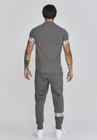 Grijze sport T-shirt met korte mouwen en een witte logoband; gecombineerd met grijze joggers met een witte logo op de boorden. Witte sneakers.