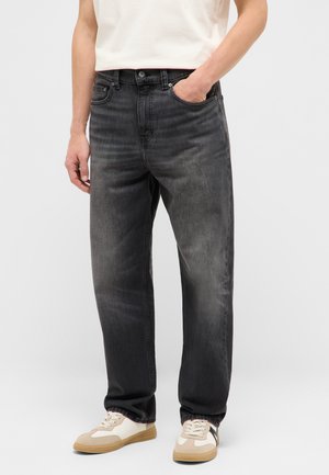 Mann trägt ausgewaschene schwarze Straight-Leg-Jeans mit weißen Sneakers und einem in die Hose gesteckten weißen T-Shirt, steht vor einem einfarbigen Hintergrund.