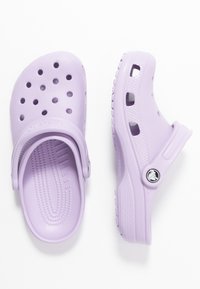 Cloșuri din plastic violet cu orificii de ventilație, curea pentru călcâie și talpă texturată. Prezintă un logo circular pe lateral.