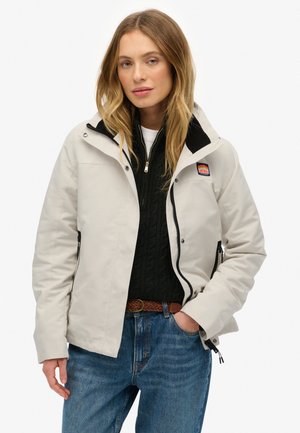 Lichtgrijze waterdichte jas met een zwarte kraag, ritszakken en een logopatch, gedragen over een donkere trui, gecombineerd met blauwe jeans.