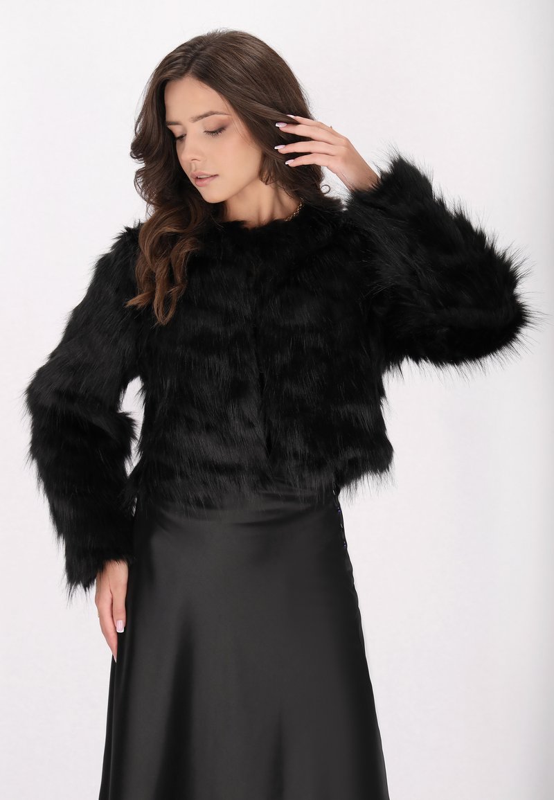 Zwarte faux fur cropped jack met lange, fluffy mouwen, voorzien van een ronde halslijn en een zachte, gestructureerde afwerking. Gedragen over een zwarte satijnen rok.