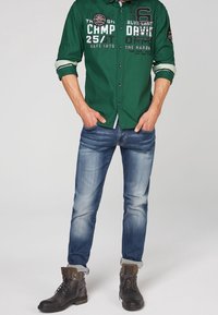 Chemise verte à manches longues avec texte imprimé, associée à un jean bleu délavé. Des bottes foncées avec les revers retroussés complètent la tenue.