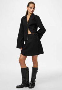 Femme portant une veste noire zippée sur le devant et une jupe portefeuille, avec des bottes en cuir noires montant jusqu'aux genoux, debout devant un fond blanc uni.