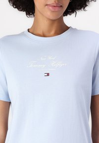 T-shirt en coton bleu clair avec un col rond, comportant un texte doré : "New York Tommy Hilfiger" et un petit logo en bas.