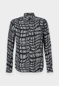 Melna krekla ar atkārtotu balto tekstu, kas satur "VERSACE" un "JEANS", garām piedurknēm un standarta apkakli. Gluda auduma tekstūra.