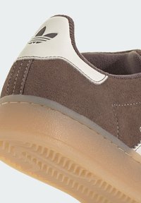 Brown suede sneaker with a white leather heel tab. Features a textured rubber sole and a small logo on the back.