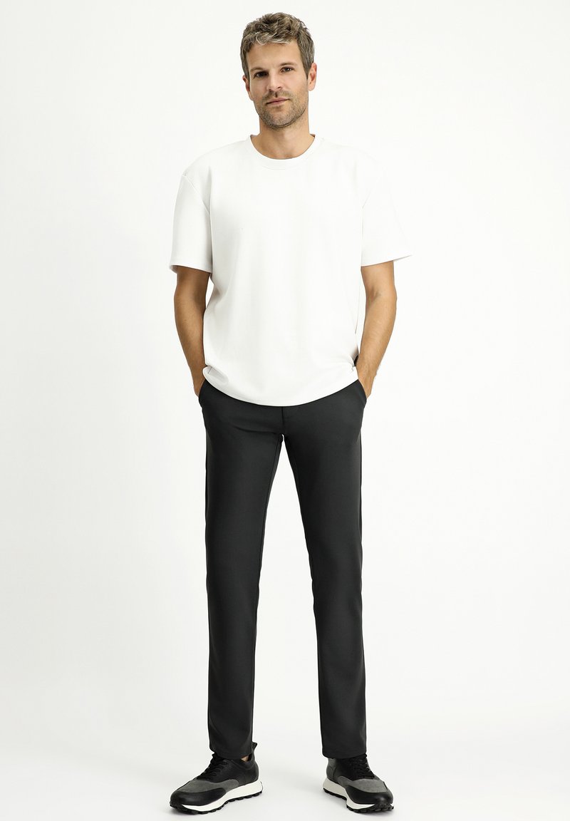 T-shirt bianca a maniche corte con scollatura rotonda, abbinata a pantaloni neri a gamba affusolata. Il modello indossa sneakers nere e grigie.