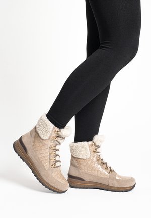 Legs in black leggings wearing beige winter boots with shearling lining, gold eyelets, and textured panels, standing on white background.