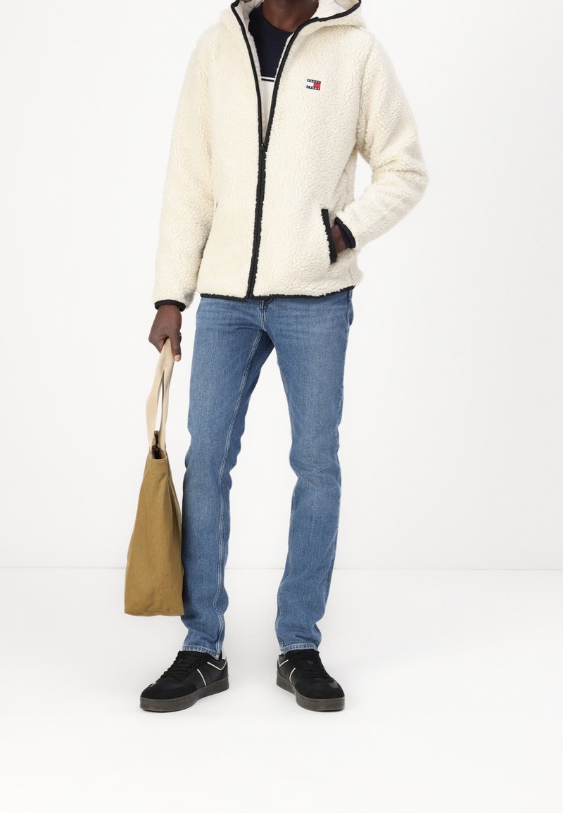 Person klädd i en vit fleecejacka, blå jeans, svarta sneakers och hållande en beige tygväska mot en vit bakgrund.