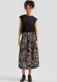 Žena v bezrukávovom čiernom topi, strední sukni s pruhovaným vzorom zebra v ružovej a čiernej farbe a čiernych členkových topánkach, stojí pred svetlosivým pozadím.