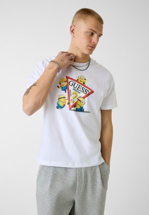 Jeune homme aux cheveux blonds courts portant un t-shirt blanc avec des Minions colorés et le logo Guess, associé à un pantalon de survêtement gris clair.