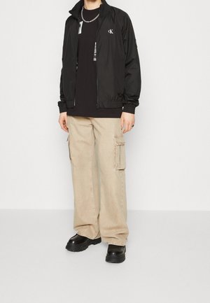 Černá zipová bunda s logem, černé tričko, béžové cargo kalhoty s kapsami a černé boty s hrubou podrážkou.