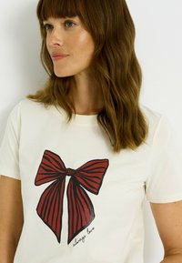 Camiseta de manga corta blanca con un gran gráfico de un lazo a rayas rojas y acabado brillante, y el texto "siempre amar" debajo.