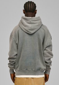 Grå pullover hættebluse med snor i huen, oversized pasform og ribbede manchetter. Blødt stof med en falmet tekstur, vist fra bagsiden.