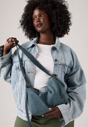 Eine Frau mit lockigem Haar trägt eine helle Jeansjacke und ein weißes Hemd. Sie hält eine blaue Jeansumhängetasche mit einem dunklen Riemen.