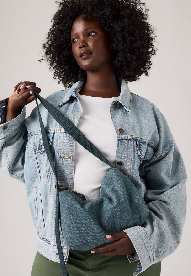 Donna con capelli ricci, che indossa una giacca di jeans chiara e una camicia bianca, tiene una borsa a tracolla di jeans blu con una tracolla scura.