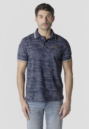 Egy férfi sötétkék mintás pólóingben, sárga csíkkal díszített gallérral és világoskék farmernadrágban, sima világos háttér előtt áll.