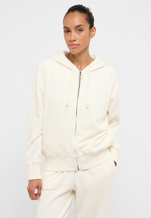 Kobieta z upiętymi włosami, ubrana w kremową bluzę zapinaną na zamek i pasujące spodnie, stoi na tle jasnego, jednolitego tła.