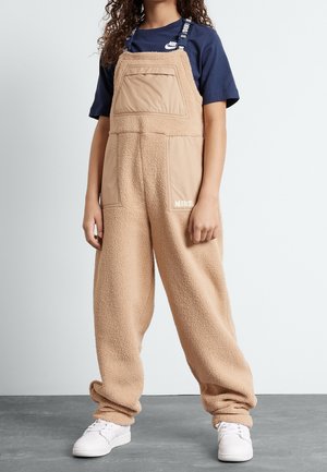 Personne portant une salopette en polaire beige avec des poches avant et des bretelles par-dessus un t-shirt Nike bleu marine et des baskets blanches, debout sur un sol gris.