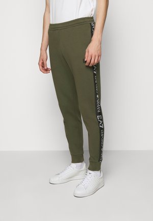 Man draagt olijfgroene joggers met zwarte zijstrepen met witte Emporio Armani EA7-logo's, witte sneakers en een wit T-shirt.