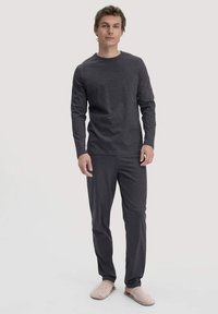 Mann, der ein dunkelgraues Langarmoberteil und passende Hose trägt, mit beigen Hausschuhen, steht vor einem schlichten hellen Hintergrund.