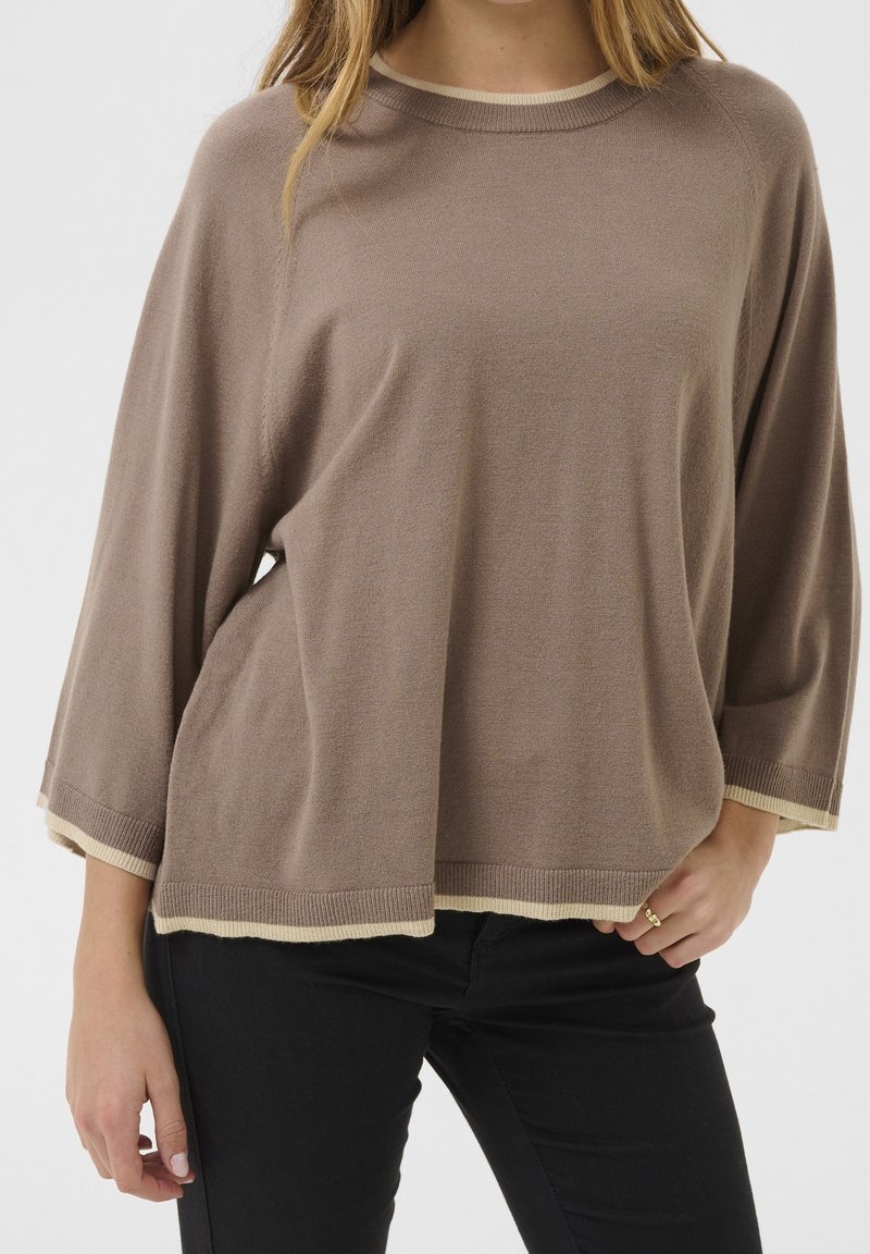 Femme portant un pull marron ample avec des bordures beiges et un pantalon noir, main posée sur la hanche, sur un fond blanc uni.