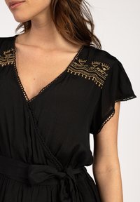 Zwarte wikkeljurk met korte mouwen, kantdetails en gouden borduursels bij de halslijn. Voorzien van een stoffen riem die om de taille is geknoopt.