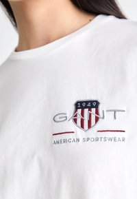 T-shirt bianca in cotone con un'emblema sul petto in rosso e blu, con "GANT" e "AMERICAN SPORTSWear" ricamati in grigio. Maniche corte.