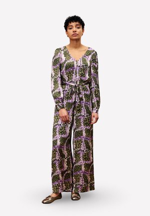 Vrouw met kort, krullend haar, gekleed in een groene en paarse gestreepte lange jumpsuit met brede benen en metalen ballerina's, staande tegen een witte achtergrond.