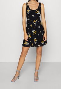 Vestido negro con flores amarillas y grises, diseño sin mangas, falda fluida. Combinado con tacones ligeros de tiras azules.