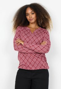 Červená košeľa s bielym geometrickým vzorom, dlhé rukávy, bez goliera a voľný strih; nosená s čiernymi nohavicami.