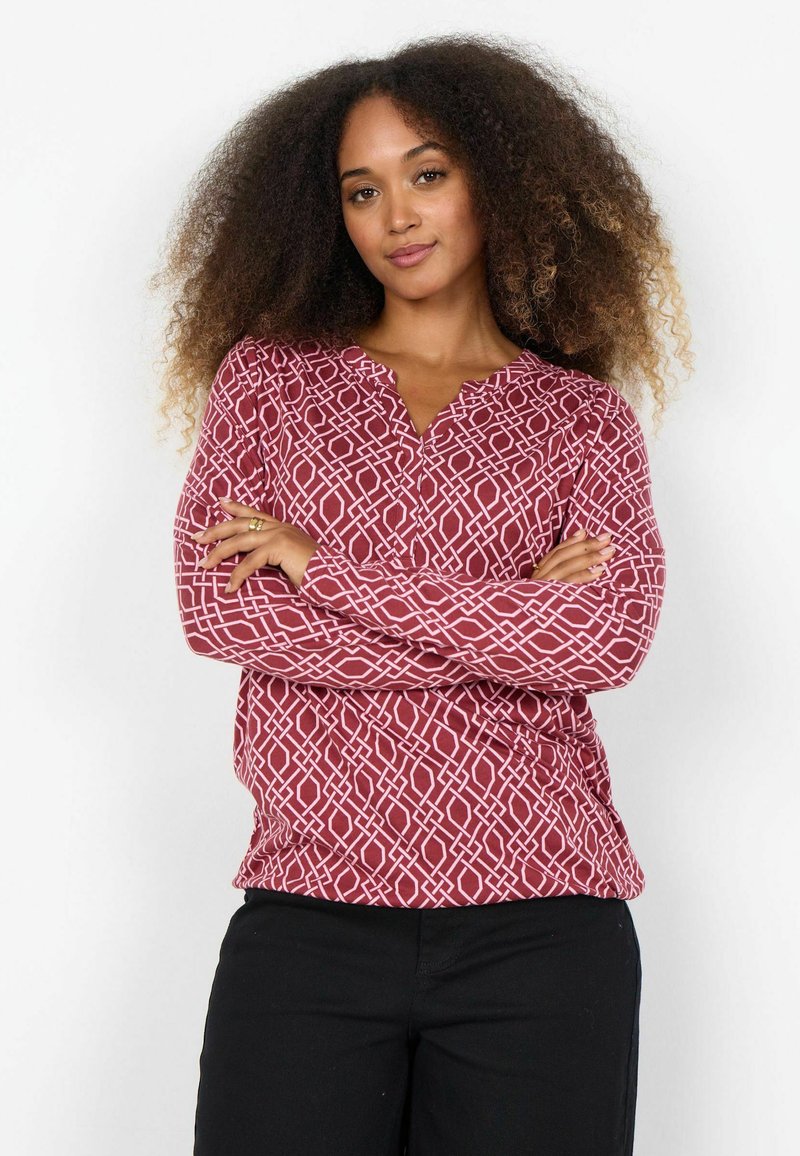 Červená košeľa s bielym geometrickým vzorom, dlhé rukávy, bez goliera a voľný strih; nosená s čiernymi nohavicami.