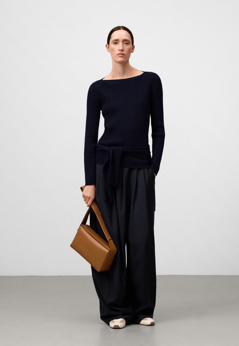 Maglione a coste blu navy con scollo largo e dettaglio a fiocco in vita, abbinato a pantaloni scuri morbidi e una borsa strutturata marrone.