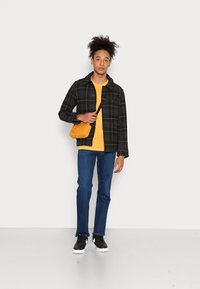 Chaqueta de franela negra y a cuadros sobre una camiseta amarilla, combinada con jeans azules y zapatillas negras. Lleva un pequeño bolso amarillo de hombro.