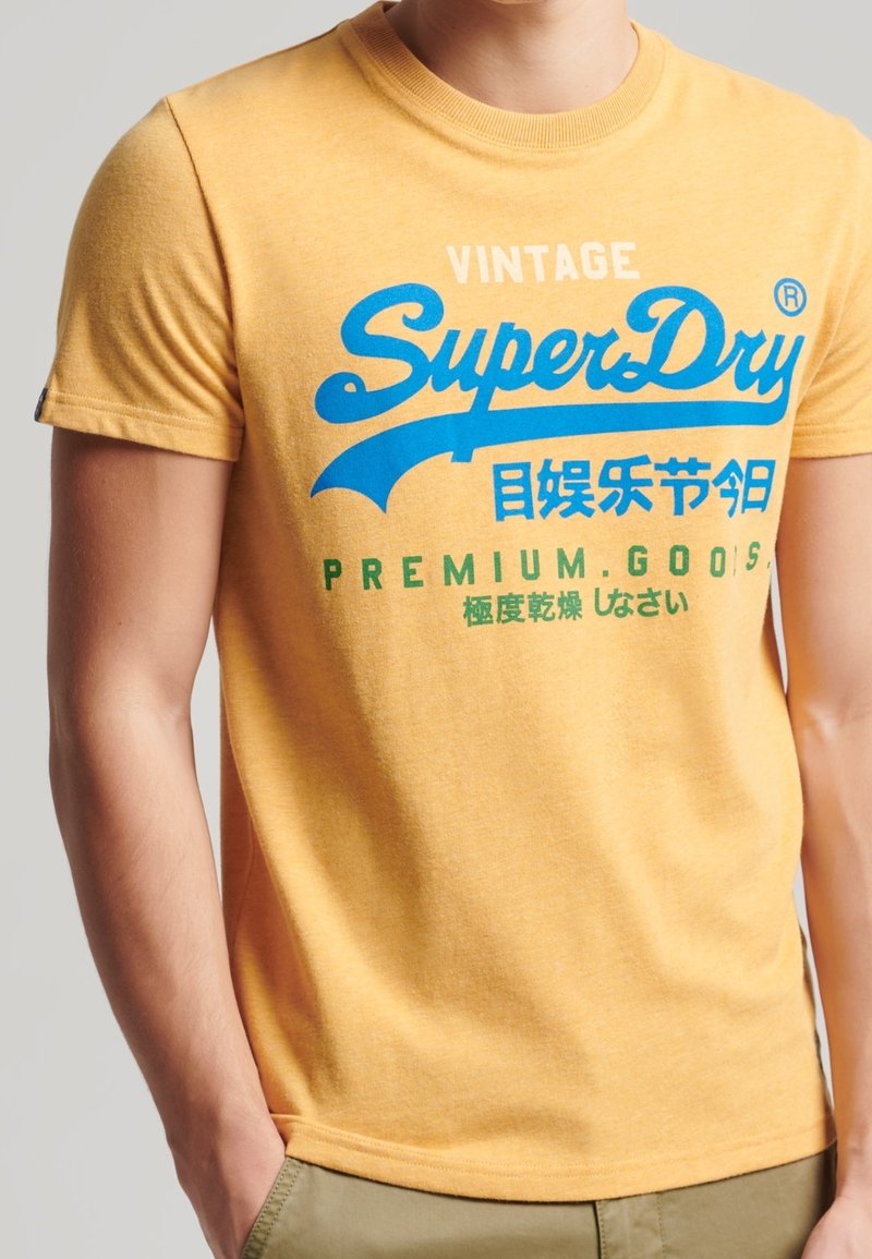 Camiseta de algodón amarilla con texto gráfico azul "SuperDry" y "PREMIUM GOODS" en verde en el frente; de manga corta y con cuello redondo.