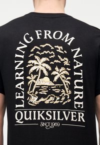Άτομο που φοράει μαύρο πουκάμισο με μπεζ τροπικό νησιωτικό σχέδιο και κείμενο "Μαθαίνοντας από τη φύση Quiksilver από το 1969" στο πίσω μέρος.