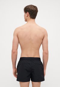Pantaloncini da mare neri con cintura elastica, texture liscia e senza motivi visibili, indossati da un modello uomo senza maglietta visto di spalle.