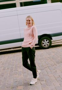 Růžový svetr s vysokým límcem a pleteným vzorem, černé cargo kalhoty a světlé tenisky. Stojící proti bílé dodávce na kostkované dlážděné cestě.