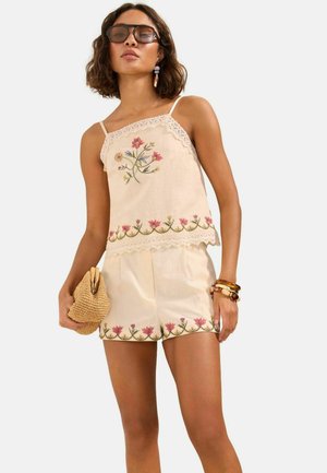 Kvinde iført beige blomsterbroderet camisole og matchende shorts, der holder en flettet clutch og bærer solbriller og armbånd.