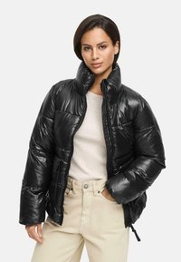 Black puffer jacket with a glossy finish, high collar, zip closure, and curved quilted pattern, paired with a light sweater and beige pants.