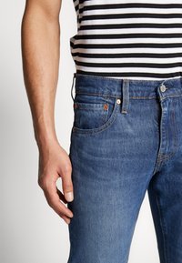 Jeans i denim med en medelblå tvätt, femfickorsdesign och kontrasterande orangea sömmar. Bärs med en svart och vit randig skjorta.