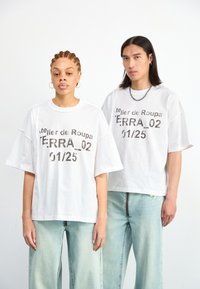 Fehér, túlméretezett pólók fémes nyomattal, amelyen az "atelier de Roupa TERRA_02 01/25" felirat szerepel, világoskék farmernadrággal párosítva.