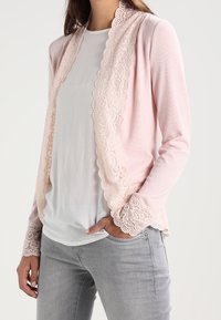 Ljusrosa kofta med spetsdetaljer längs kanterna och ärmarna, med en texturerad ribbad tyg. Buren över en vit topp.