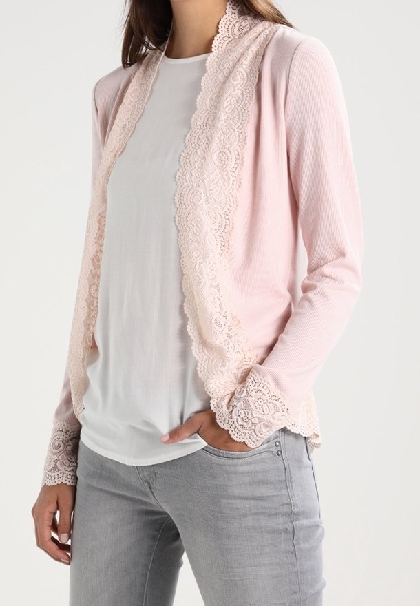 Ljusrosa kofta med spetsdetaljer längs kanterna och ärmarna, med en texturerad ribbad tyg. Buren över en vit topp.