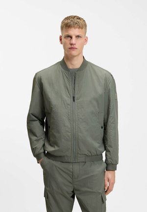 Joven con chaqueta bomber verde oliva y pantalones a juego, de pie con las manos en los bolsillos frente a un fondo blanco liso.