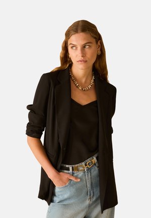 Donna con capelli mossi indossa blazer nero e top, jeans azzurri chiari con cintura con fibbia dorata, collana con conchiglia e orecchini a cerchio dorati, guarda di lato.