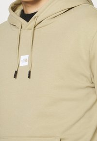 Hoodie beige chiaro realizzato in morbido tessuto, con tasca frontale, cordini regolabili e una piccola etichetta del logo.